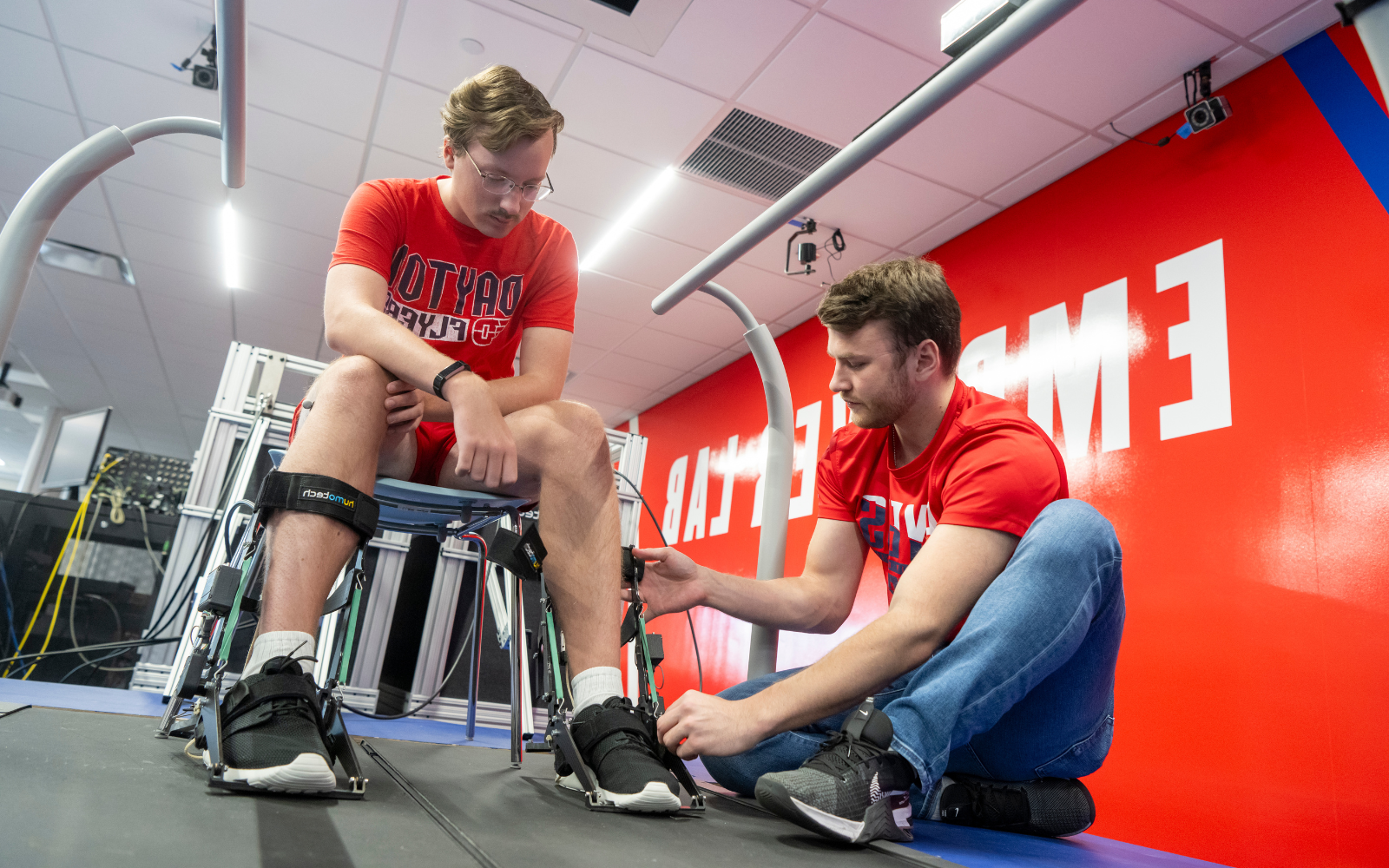 A male student helping a male put on leg braces while sitting in a chair.